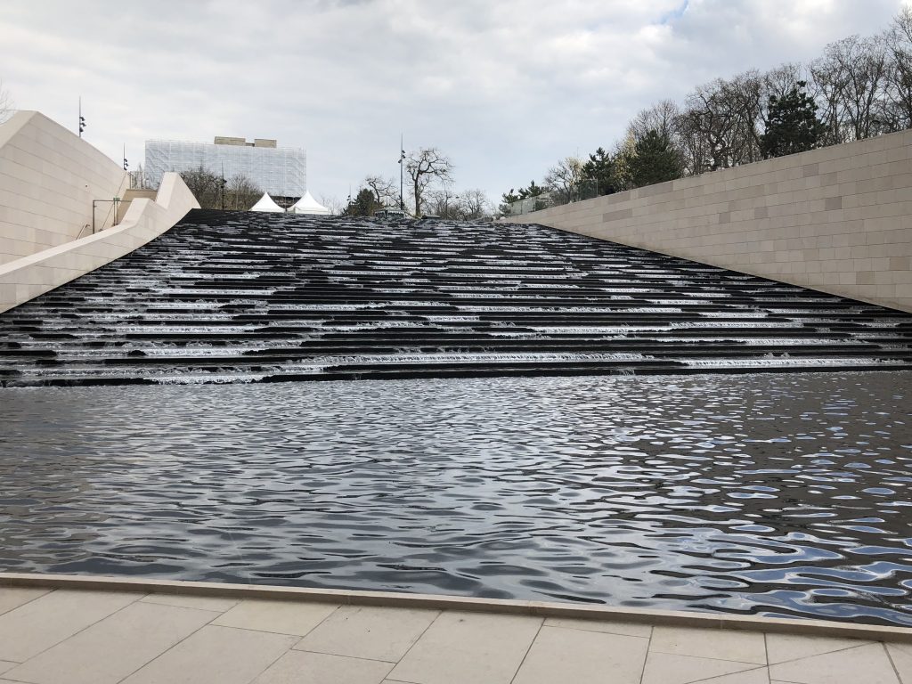 An outside waterfall cascades via steps into a basin outside the auditorium of the Louis Vuitton Foundation building. 
